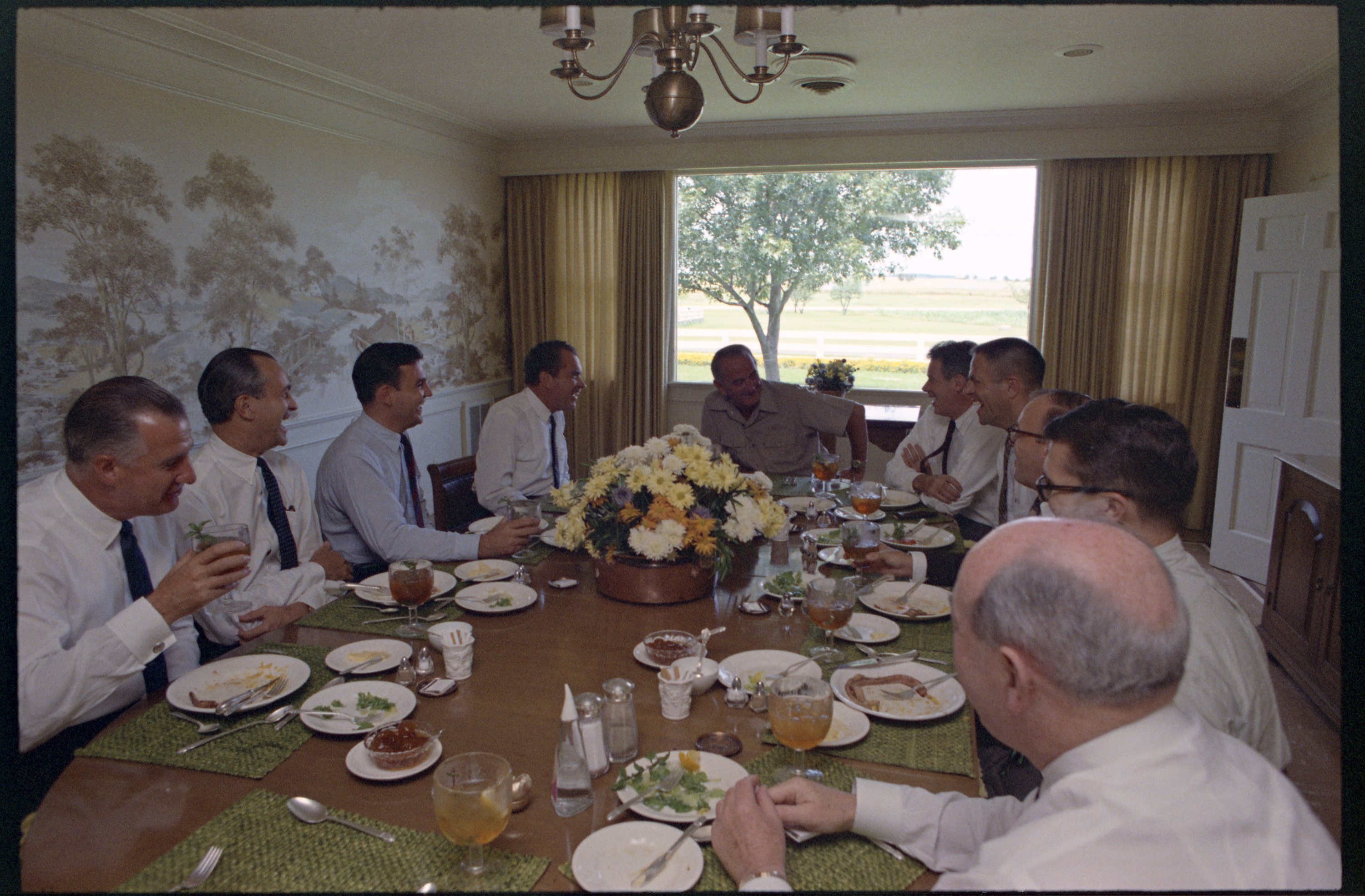 A group of men in shirtsleeves and ties sit around an elliptical table, all looking at President Lyndon B. Johnson, center, in khaki leisurewear, and laughing at something he has said.